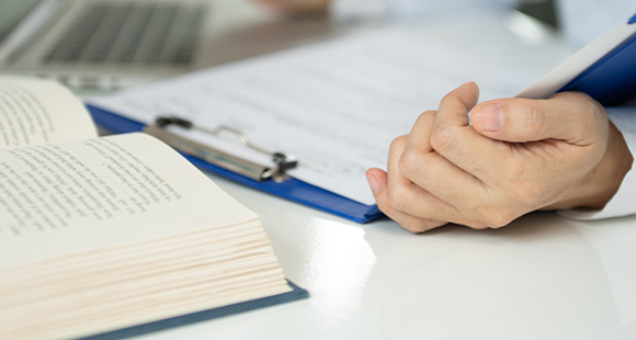 person's hands holding a clipboard with documents beside an open book and a laptop on a desk six essential study materials